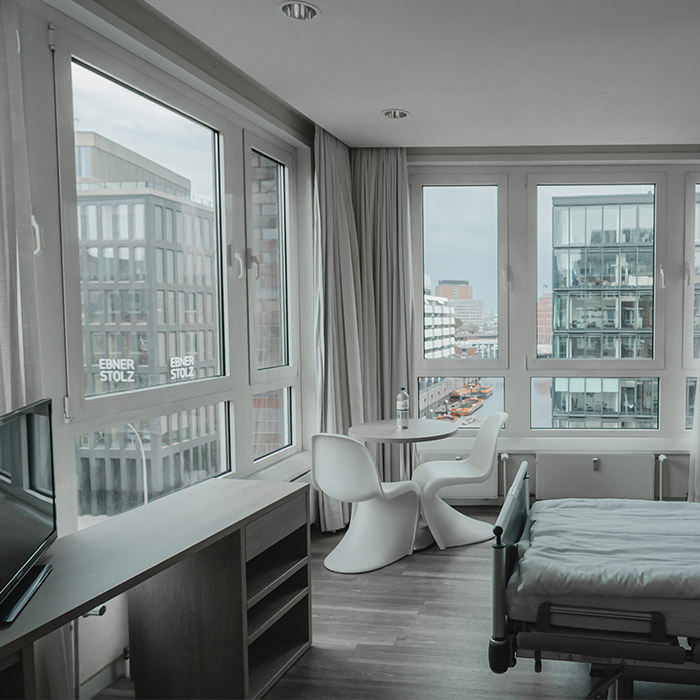 Modernes Patientenzimmer mit Schreibtisch, Bett, Fernseher und Tisch und großen Fenstern mit Ausblick auf benachbarte Gebäude. 