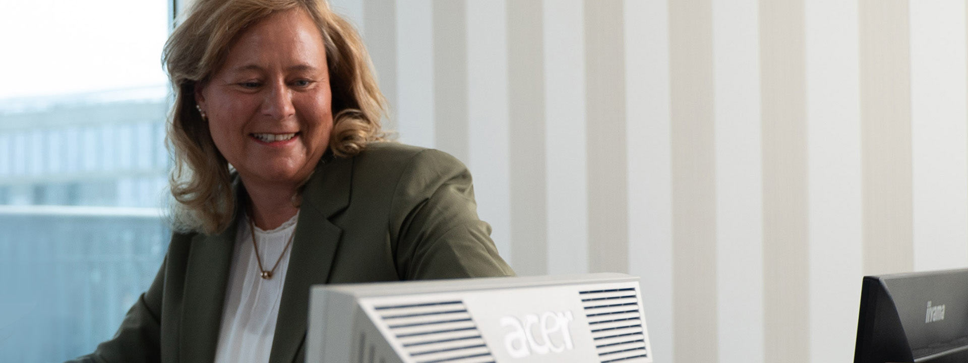 Mitarbeiterin der Privatpraxis für Orthopädie mit Blick auf einen Bildschirm am Schreibtisch vor großem Fenster sitzend
